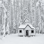Simple black and white vector line art of a cozy cabin surrounded by pine trees and snow, designed as a children's printable coloring page.
